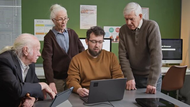 Medium shot of male teacher demonstrating tasks on laptop screen to group of senior people attending computer literacy courses in school classroom