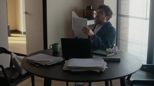 High angle of mature Caucasian male writer wearing robe sitting at table in living room, proofreading manuscript