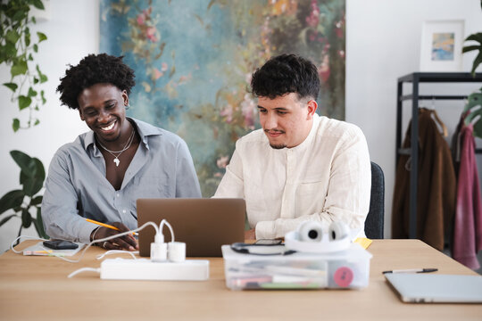Two young men collaborate on a project at a desk, one smiling while writing and the other focused on a laptop.