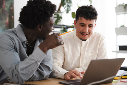 Two young men collaborate on a project, one pointing with a pencil while the other uses a laptop, sharing ideas and working together.