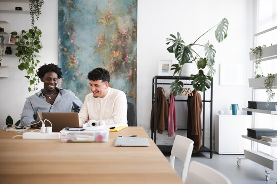 Two young men collaborate on a laptop at a wooden desk in a bright, modern office space filled with plants and artwork.