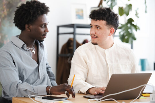 Two young men collaborate on a project at a desk, one using a laptop while the other holds a pencil, engaged in discussion.