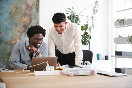 Two men collaborate on a project at a desk, one using a laptop while the other leans in to discuss.