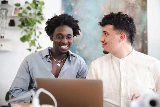 Two young men collaborate on a project using a laptop, sharing ideas and smiling in a bright, modern office space with plants.