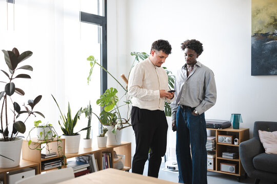 Two men stand in a bright room filled with plants, one holding a smartphone and showing it to the other.