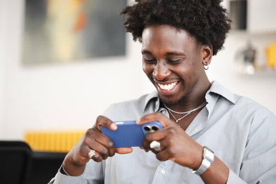 A young Black man smiles while looking at his phone. He is wearing a light blue collared shirt and silver jewelry.