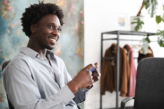 A young man smiles while holding a small electronic device in his hands. He is seated in a chair in a room with a colorful mural on the wall.
