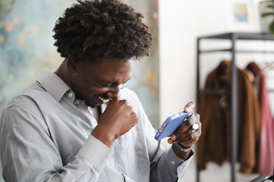 A young Black man in a light blue collared shirt laughs heartily while looking at his phone.