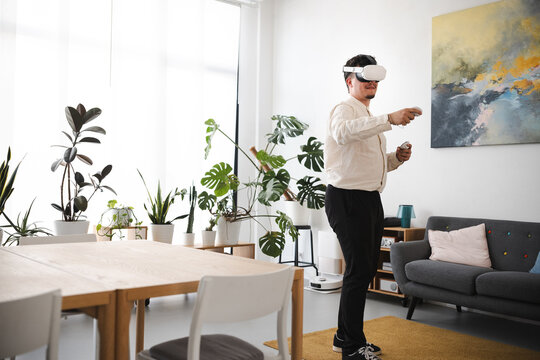 A man wearing a virtual reality headset and holding controllers interacts with a digital world in a modern, plant-filled living room.