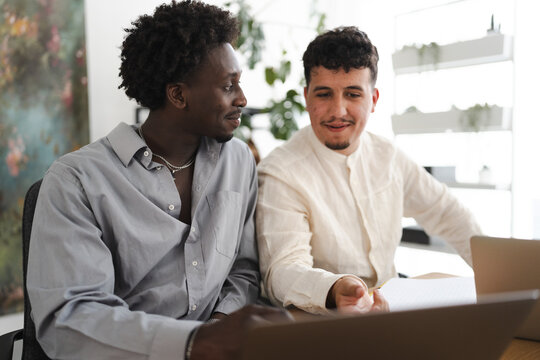 Two men collaborate on a project, looking at a laptop and papers in a bright, modern office setting.
