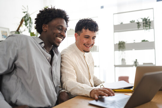 Two men collaborate on a project, smiling as they work together on a laptop and review documents at a wooden desk in a bright, modern office space.