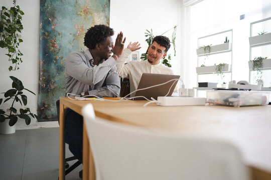 Two men give each other a high five while working together at a desk with a laptop and plants in a bright, modern office space.