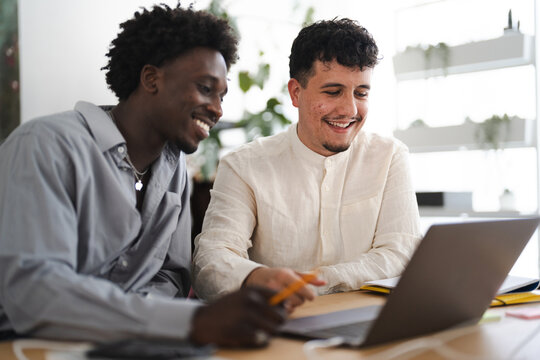 Two young men collaborate on a project, smiling as they review information on a laptop at a desk.