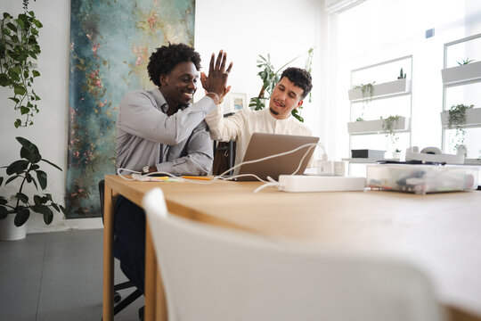 Two men celebrate a successful project with a high five while working together at a desk with a laptop and plants in a bright office.