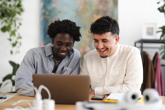 Two young men collaborate on a project, smiling as they look at a laptop screen together at a desk.