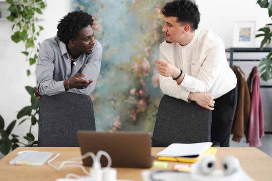 Two men are engaged in a conversation while leaning on chairs in a modern office setting.