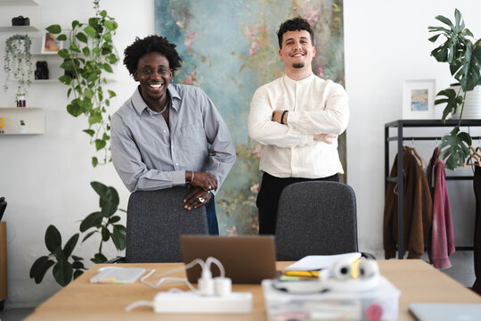 Two young men smile at the camera in a modern office space. One leans on a chair, the other stands with arms crossed.