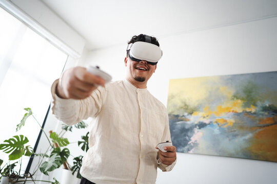 A man wearing a virtual reality headset and holding controllers interacts with a digital world, experiencing immersive technology in a modern room.