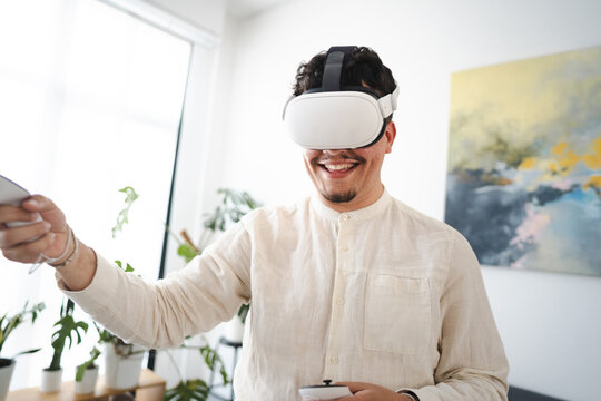 A man wearing a virtual reality headset smiles as he holds controllers, engaging with immersive digital content in a bright, modern room.