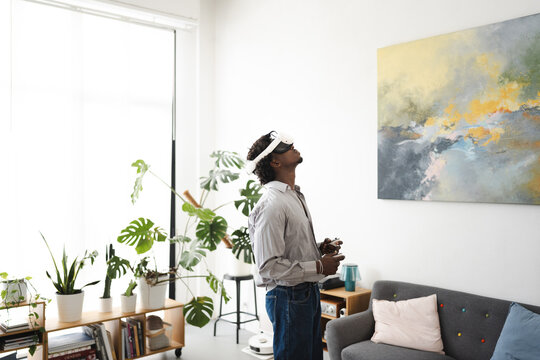 A man wearing a virtual reality headset and holding controllers looks up in awe, experiencing immersive technology in a modern living room.
