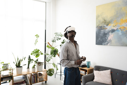 A young man wearing a virtual reality headset and holding controllers immerses himself in a digital world while standing in a modern living room.