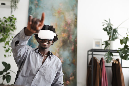 A young man wearing a virtual reality headset smiles as he reaches out his hand, engaging with an immersive digital experience in a modern, plant-filled room.