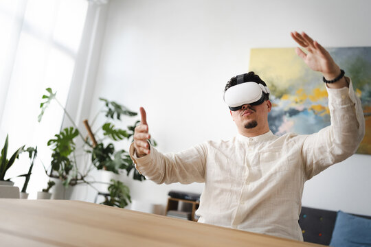 A man wearing a virtual reality headset interacts with a digital world, his hands outstretched in a modern, bright room with plants.
