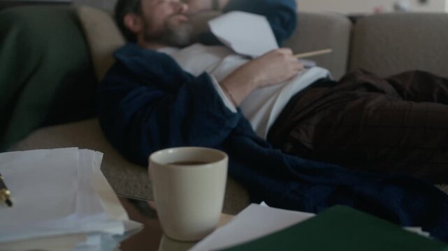Middle aged Caucasian male author taking nap on sofa while proofreading novel draft, mugs, bowl with snacks and papers visible on coffee table in foreground