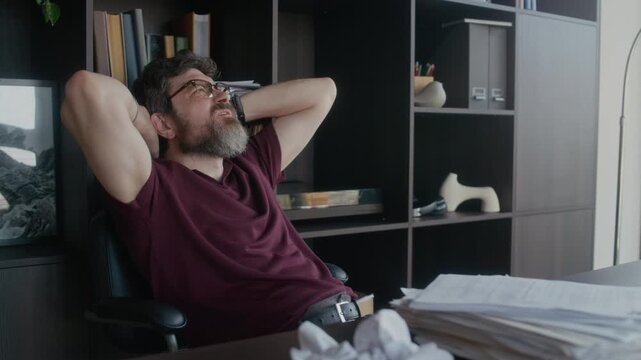 Medium shot of frustrated mature adult Caucasian male writer experiencing creative block sitting in home office, grumbling and rubbing nose bridge, crumpled draft visible on table