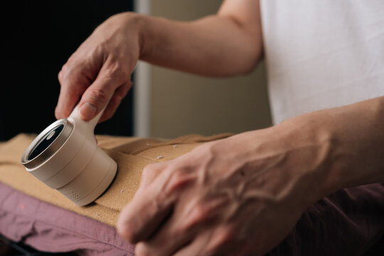 Close-up cropped shot of unrecognizable person using electric fabric shaver to remove pilling and lint from brown sweatshirt, enhancing garment care and maintaining textile quality in home.