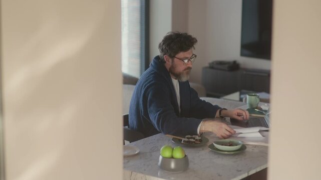 Side view of mature adult Caucasian male writer with beard on face sitting at kitchen counter working on new novel, copy space