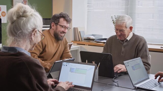 Group of senior people enjoying computer literacy class and smiling happily while using laptops at desk in school classroom with caring male teacher laughing