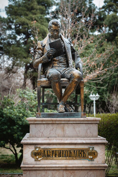 Statue of A. S. Griboedov in Park, Sitting and Reading a Book, Surrounded by Trees and Plants, Well-Maintained Area, Warm Day in Early Spring