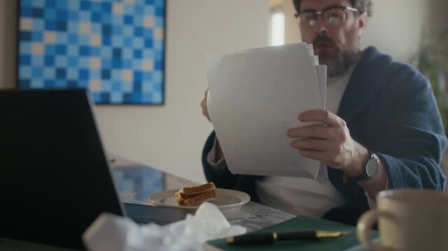 Mature Caucasian male writer wearing robe drinking coffee while reading draft in living room at home, sitting at table with laptop, plate with sandwich and crumpled papers on it