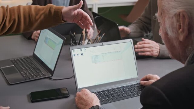 Over shoulder close up shot of senior man using laptop in computer literacy class and asking questions while unrecognizable teacher explaining task