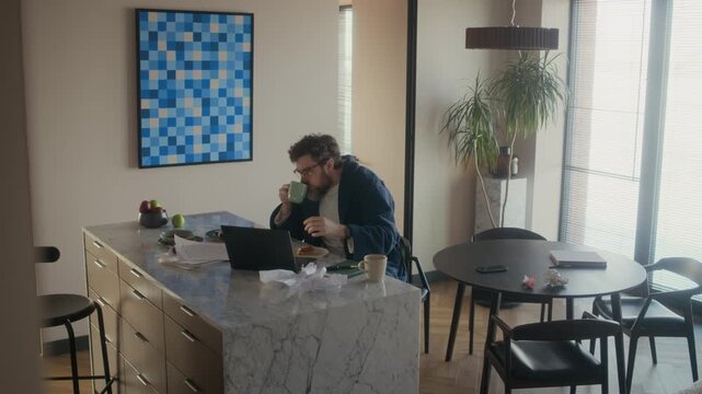 Wide shot of Caucasian male writer wearing robe eating sandwich for lunch, wiping hands and getting back to work with novel draft