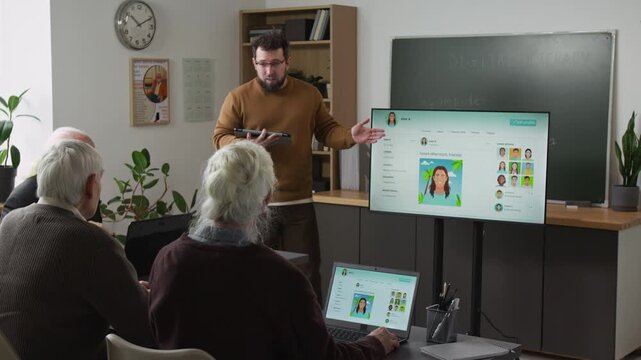 Male teacher giving lecture in computer class for seniors and pointing at digital screen with personal social media page explaining online communication to group of adult students, copy space