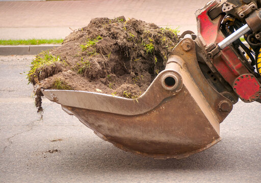 Close-up of Excavator Bucket Holding Soil and Grass; Conceptual Image for Civil Engineering, Site Preparation, and Construction Progress