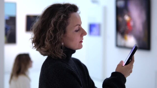 Woman with curly hair standing in a modern art gallery, attentively using her smartphone while surrounded by paintings and other visitors, blending technology with cultural appreciation
