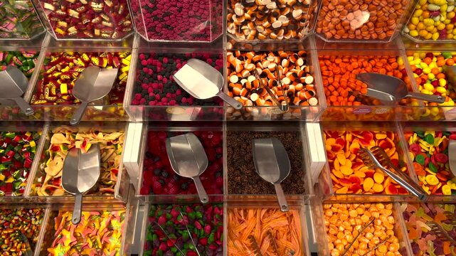 Colorful selection of assorted gummy candies and sweets displayed in bulk bins at a market. Metal scoops and tongs are ready for customers to pick their favorite sugary confections