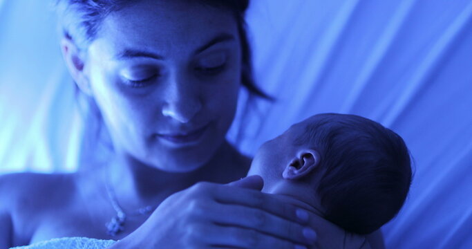 Mother holding newborn baby at hospital next to violet phototherapy lamp, real life affection and love