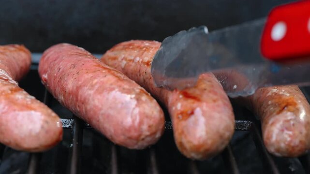 View of raw sausages turning over while grilling on barbecue with open flame. Close up of juicy meat cooking on grill, outdoor food and summer barbecue