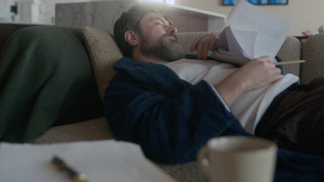 Tired mature Caucasian male novelist holding pencil in hand sleeping on sofa in living room, pile of papers, pen and mug visible in foreground