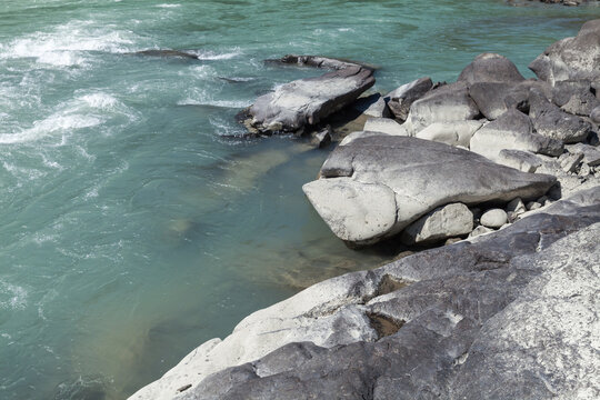 Smooth granite boulders line a turquoise mountain river, where gentle rapids and clear water flow around weathered rocks, creating a serene natural scene ideal for outdoor and travel themes