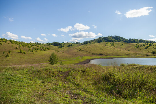 Expansive summer countryside with rolling hills, a small reflective pond and scattered trees under a bright blue sky with white clouds. Serene rural meadow scene ideal for nature, travel, and outdoor 