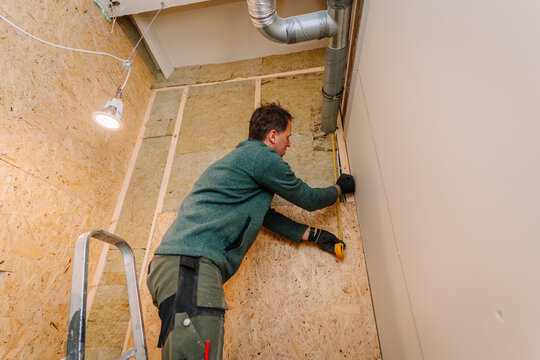 Male construction worker measures wall paneling while standing on a ladder in a room with wooden wall and exposed ductwork