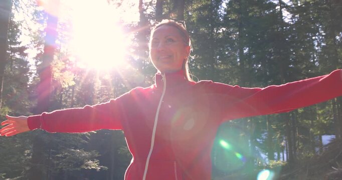 Young woman enjoys at beautiful sunset in pine forest mountain, slow motion, closeup of happy woman spreads her arms and bright sun, branches of the pine trees, season travel nature idyllic