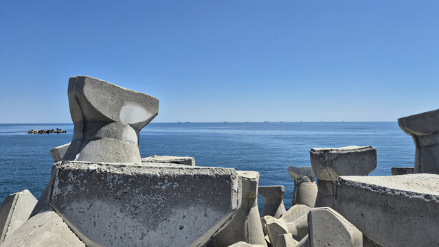 Large concrete tetrapods line the coast, serving as a breakwater against the calm blue sea. Ships are visible on the distant horizon under a clear sky, highlighting coastal engineering.