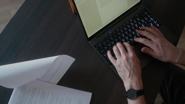 From above view of unrecognizable male author hands writing novel on laptop while sitting at desk with printed draft, notebooks and pen on it
