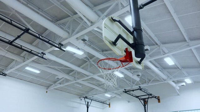 Basketball hoop and backboard inside an indoor gymnasium with bright lights and high ceiling. Sports facility interior with empty court and professional equipment.
